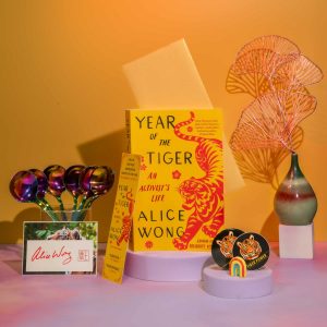 Image description: Photo with a tangerine background with a pink border at the bottom with a copy of Year of the Tiger paperback in the center. The left in a glass cup is a bunch of rainbow dipper spoons from @umeshiso_ with a signed tiger bookplate by Alice Wong in front of the cup. A tiger bookmark positioned to the left of the book. On the right is a multicolored pear-shaped vase holding gold ginkgo leaf decorations. In the front is one round tiger sticker and one round tiger button with a small rainbow decor standing upright. Photo credit: @ziru.mo (on IG)