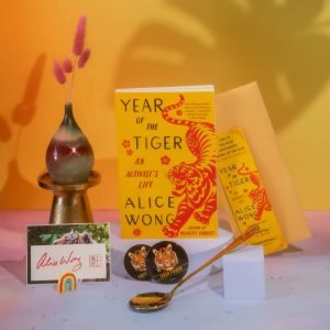 Image description: Photo with a tangerine background with a pink border at the bottom with a copy of Year of the Tiger paperback in the center. A tiger bookmark positioned to the right of the book. On the left is a multicolored pear-shaped vase holding pink floral decorations. A signed tiger bookplate by Alice Wong in front of the vase with a small rainbow decor standing upright. In the center is one round tiger sticker and one round tiger button with a gold dipper spoon from @umeshiso_ positioned horizontally at an angle. A tiger bookmark positioned to the right of the book. Photo credit: @ziru.mo (on IG)