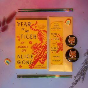 Image description: Photo with a purple-pink background with a copy of Year of the Tiger paperback in the center. Above and below the book are a pair of rainbow chopsticks from @umeshiso_ (on IG). On the right of the book is a tiger bookmark and is one round tiger sticker and one round tiger button. Floral decorations and a small rainbow decor are placed around the edges. Photo credit: @ziru.mo (on IG)